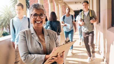 female teacher smiles with clipboard as students walk by