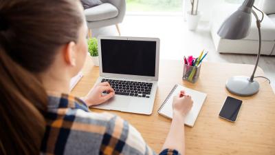 woman sits at computer desk with laptop and notepad