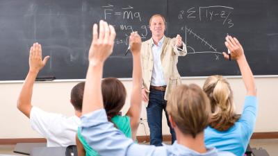 students raise hands in classroom with teacher and chalkboard