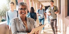 female teacher smiles with clipboard as students walk by