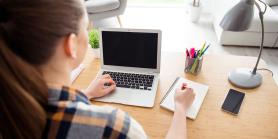 woman sits at computer desk with laptop and notepad