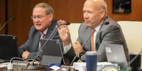 two men seated at conference table with microphones